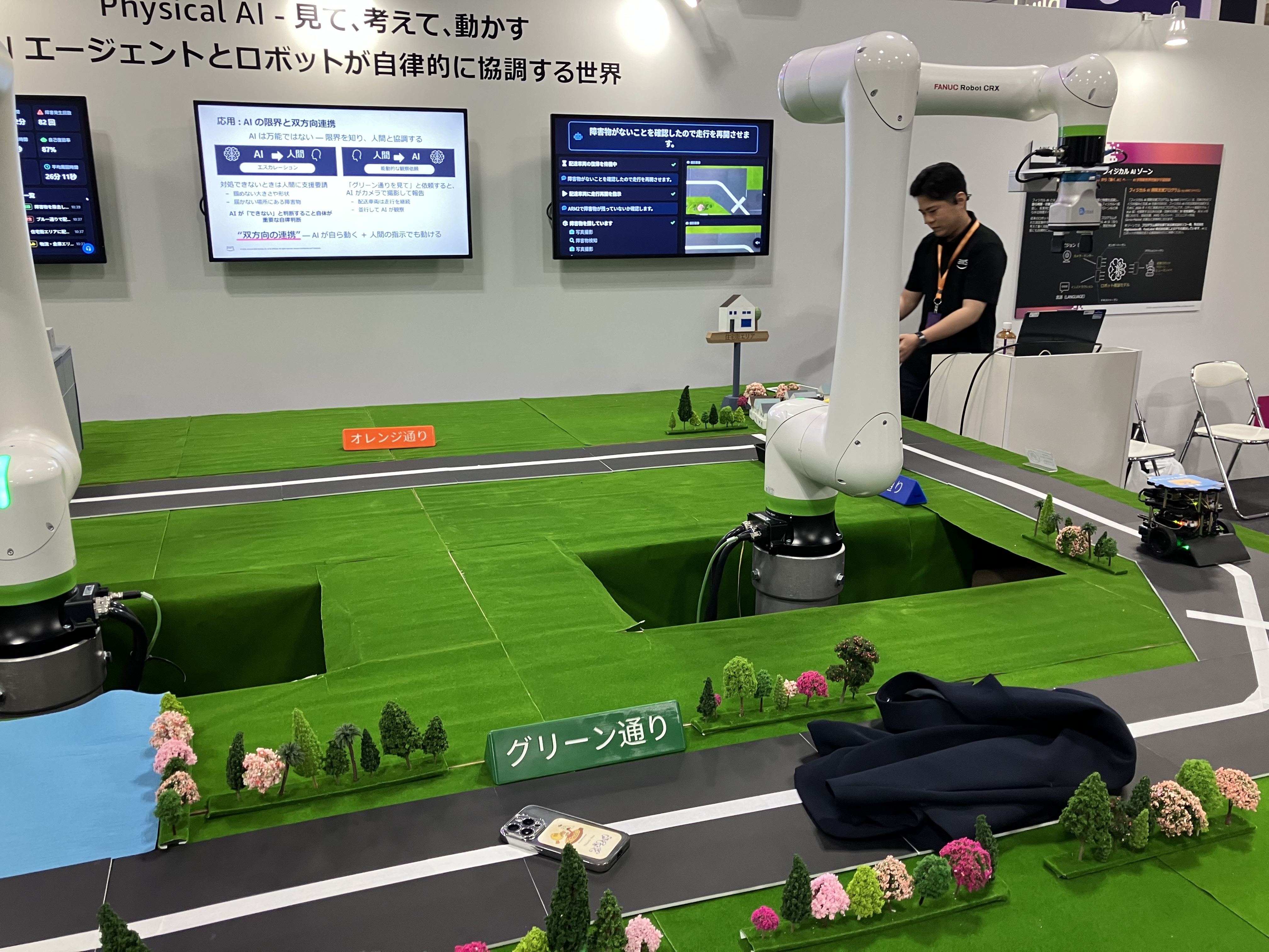道路の上に来場者のスマートフォンや黒い衣類が障害物として置かれた様子。2台のロボットアームがグリーン通りを調査する